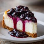 A close-up of a rich slice of blueberry cheesecake with a thick graham cracker crust and dripping blueberry topping.