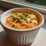 Close-up of a small white ramekin filled with bright orange remoulade sauce, garnished with chopped green onions.