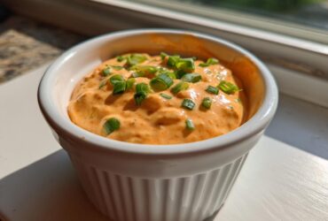 Close-up of a small white ramekin filled with bright orange remoulade sauce, garnished with chopped green onions.