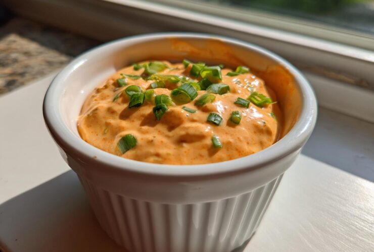 Close-up of a small white ramekin filled with bright orange remoulade sauce, garnished with chopped green onions.