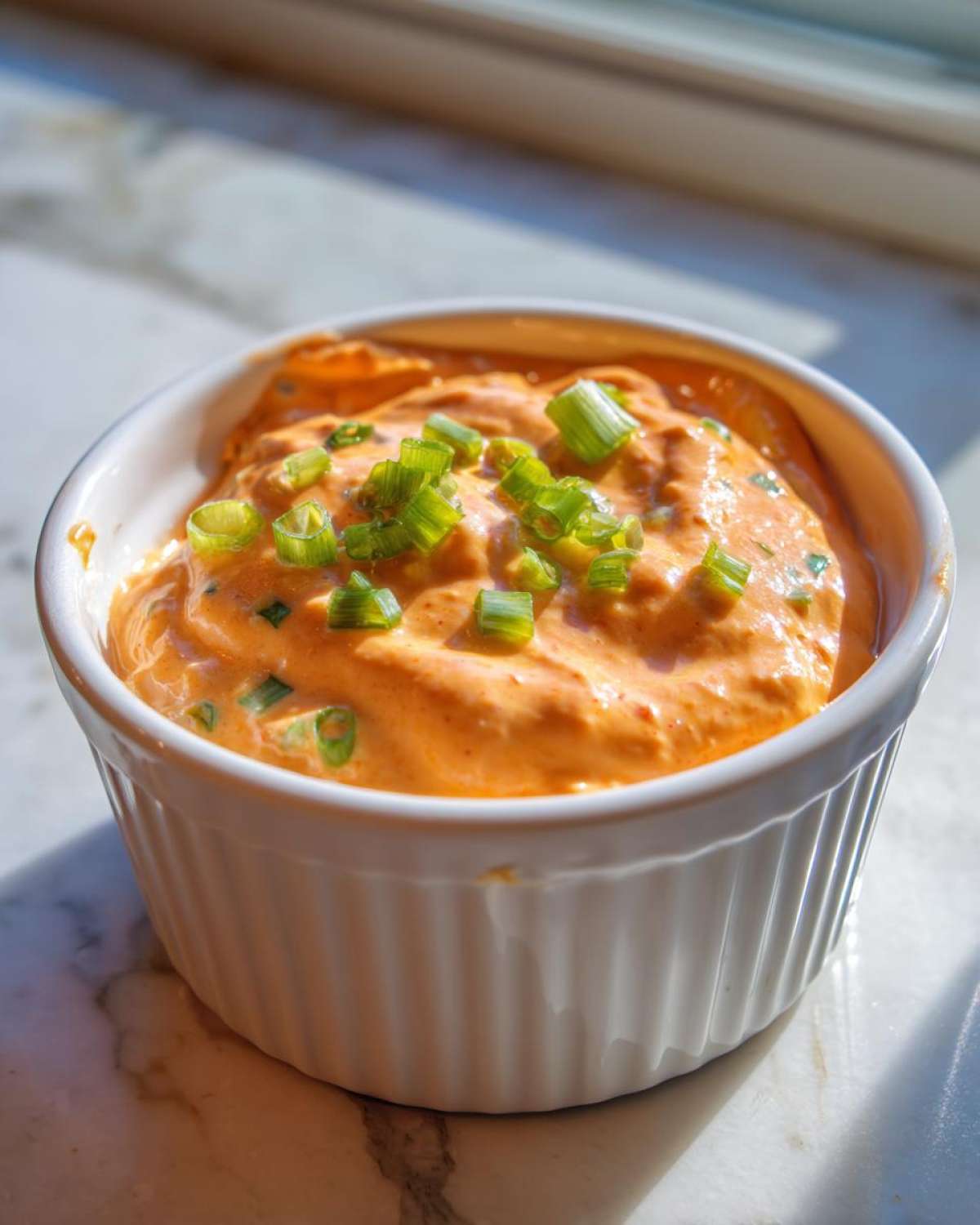 Close-up of creamy, orange-hued remoulade sauce topped with fresh green onions in a white bowl.