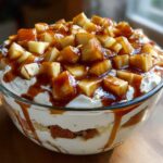 A close-up of a glass bowl filled with a layered caramel apple trifle, topped with whipped cream and caramel-coated apple chunks.
