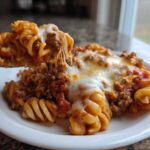A spoonful of cheesy pizza casserole with rotini pasta and meat sauce being lifted from a white plate.