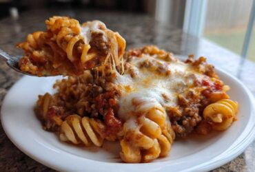 A spoonful of cheesy pizza casserole with rotini pasta and meat sauce being lifted from a white plate.