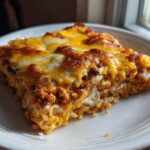 A close-up, sunlit serving of cheesy burrito bake layered with rice and ground meat mixture on a white plate.