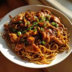 A close-up of a bowl filled with glistening chicken chow mein recipe, topped with fresh green onions.