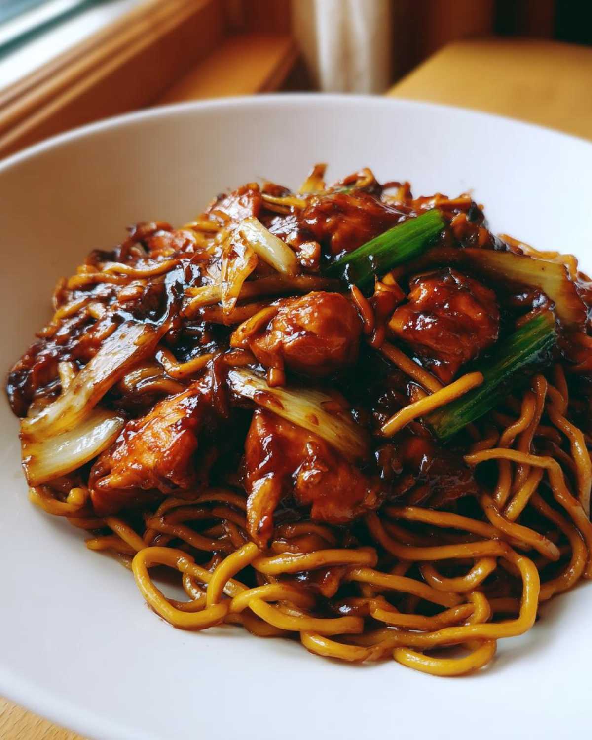 A close-up of a white bowl filled with glossy, saucy chicken chow mein recipe featuring noodles, chicken pieces, and green onions.