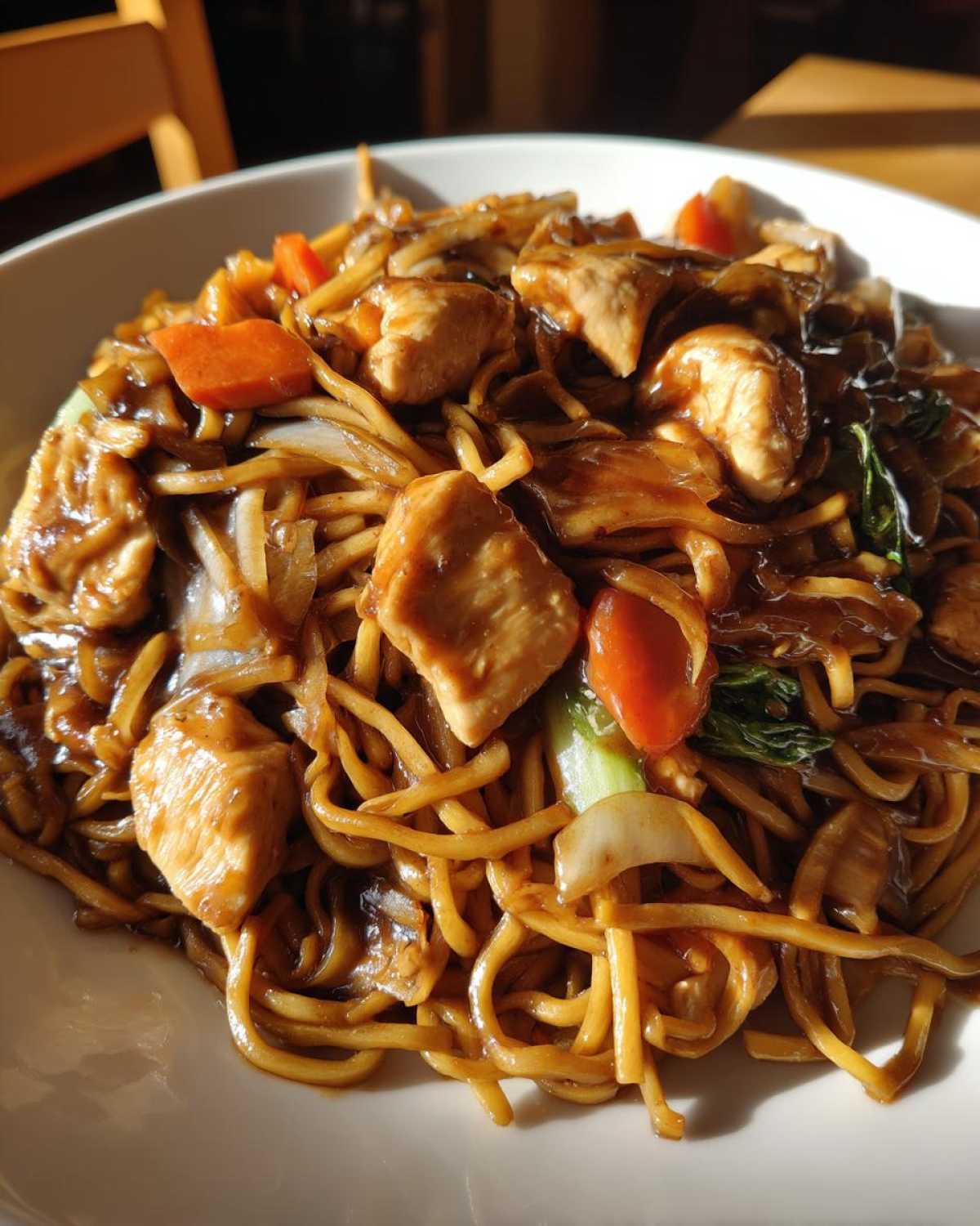 Close-up of a white bowl filled with savory chicken chow mein recipe, featuring noodles, chicken pieces, carrots, and bok choy.