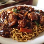 Close-up of a white plate featuring saucy chicken pieces and vegetables served over egg noodles in a chicken chow mein recipe.