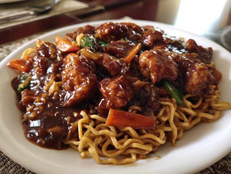 Close-up of a white plate featuring saucy chicken pieces and vegetables served over egg noodles in a chicken chow mein recipe.