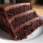A decadent slice of three-layer chocolate cake recipe covered in thick chocolate frosting, sitting on a white plate.