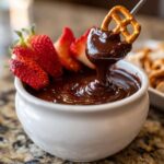 A pretzel being dipped into rich, melted chocolate fondue, with fresh strawberries nearby.