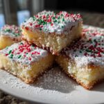 Close-up of stacked christmas gooey butter cookies squares dusted with powdered sugar and red/green sprinkles.