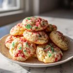 A stack of soft, buttery christmas sprinkle cookies topped with red and green holiday nonpareils on a white plate.