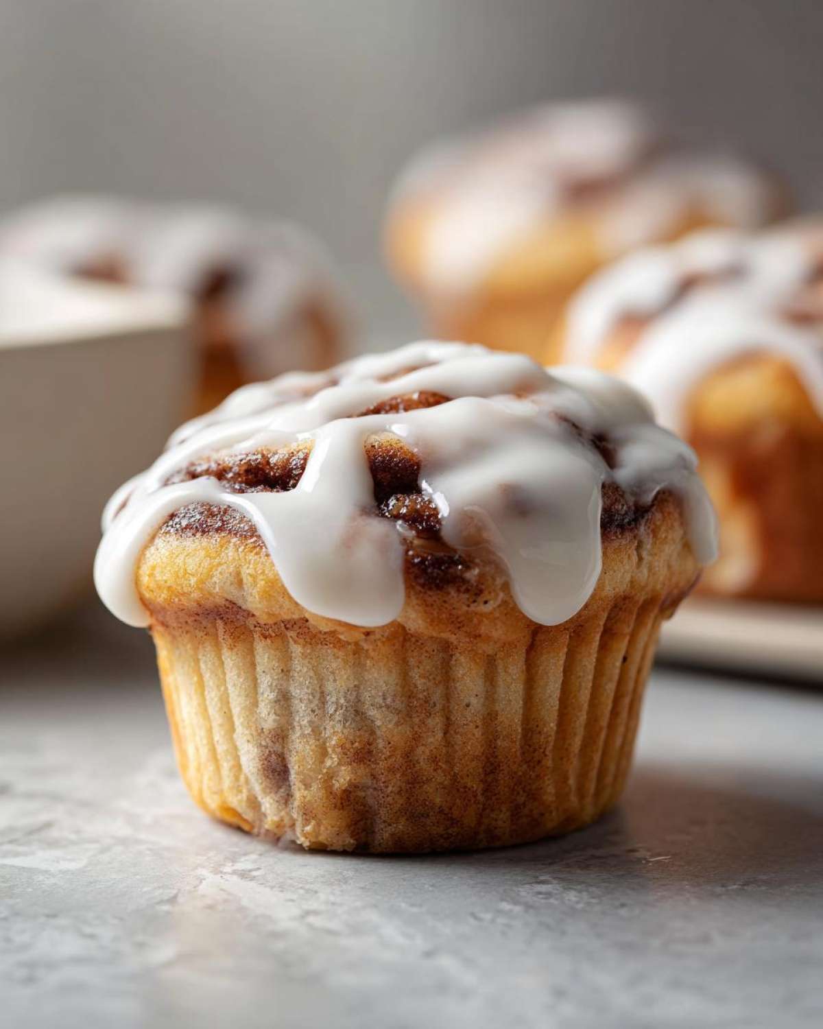 A close-up of a freshly baked cinnamon roll muffin topped with thick white icing, with more muffins blurred in the background.