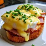 Close-up of classic eggs benedict with rich hollandaise sauce and chives on toasted bread.
