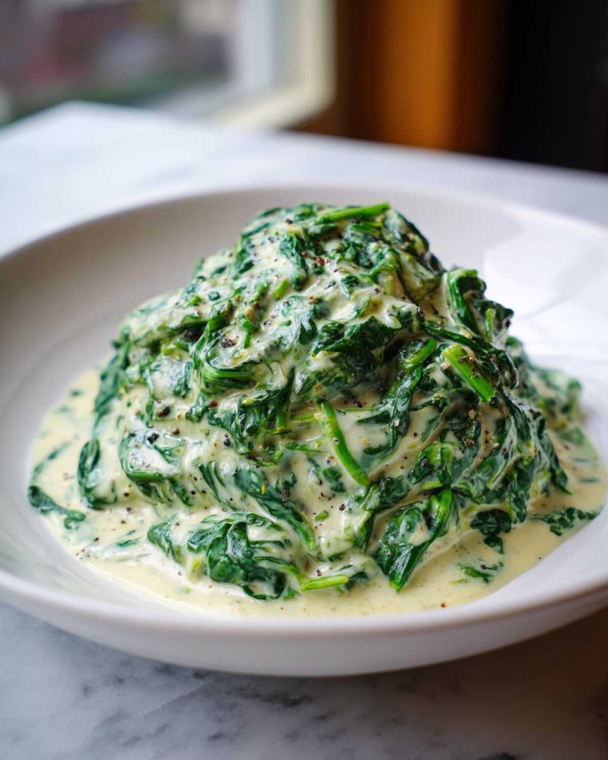 A generous serving of rich creamed spinach recipe piled high in a white bowl, seasoned with black pepper.