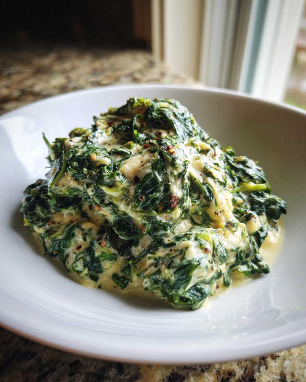 A close-up of a generous serving of rich creamed spinach recipe in a white bowl, seasoned with pepper.