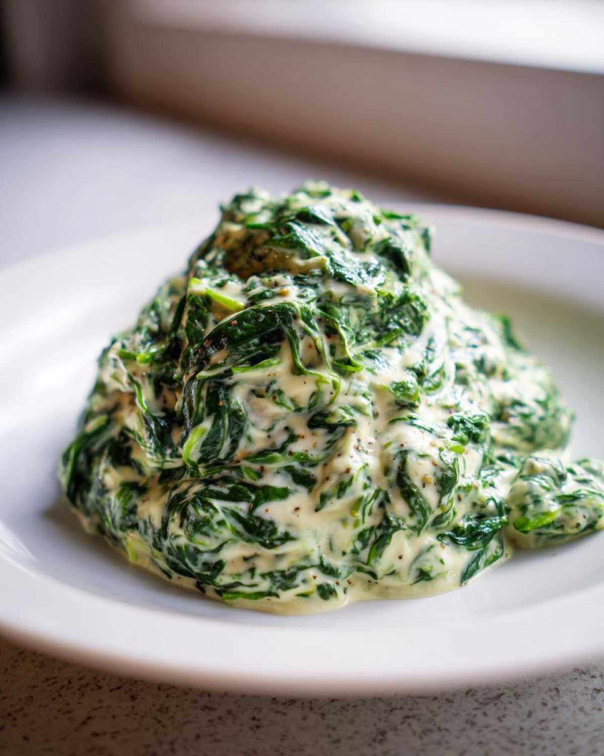 A mound of rich, homemade creamed spinach recipe served on a white plate, seasoned with pepper.