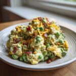 A close-up of creamy pea salad mixed with a dressing, topped with shredded cheddar cheese and crispy bacon bits.