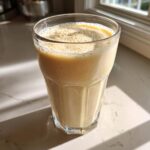 A tall glass filled with a creamy, light-colored banana smoothie, sitting on a sunny kitchen counter.
