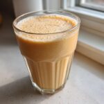 A close-up of a creamy, frothy banana smoothie served in a textured glass, sitting on a light countertop near a window.