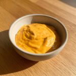 A close-up of thick, vibrant orange beer cheese dip served in a small, light-colored ceramic bowl on a wooden table.