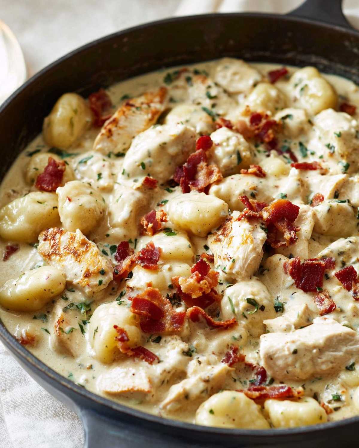 Close-up of creamy crack chicken gnocchi dish in a black skillet, topped with bacon bits.