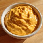 Close-up of thick, creamy orange beer cheese dip served in a small white bowl on a wooden surface.