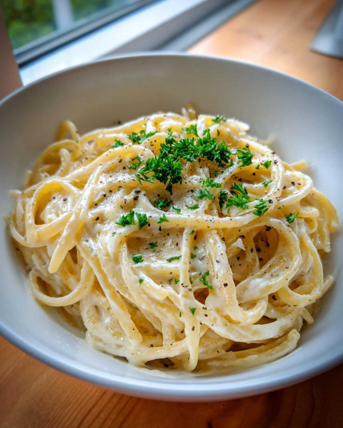 Close-up of creamy parmesan pasta tossed in a white bowl, topped with fresh parsley and black pepper.