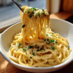 A fork lifts creamy strands of parmesan pasta coated in sauce and topped with grated cheese and parsley.