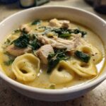 A close-up of a white bowl filled with creamy tortellini soup, featuring chunks of chicken, wilted spinach, and grated Parmesan cheese.
