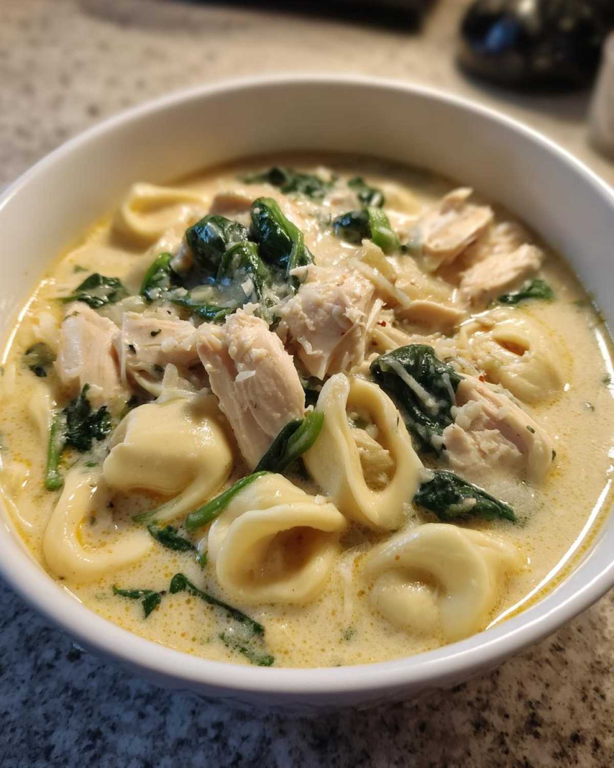 A close-up view of a bowl filled with creamy tortellini soup, featuring chunks of chicken, wilted spinach, and grated cheese.