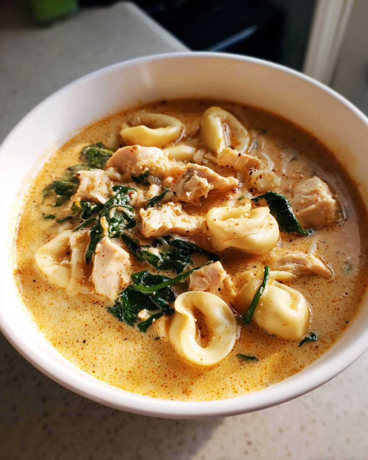 A close-up view of a white bowl filled with creamy tortellini soup, featuring chunks of chicken, wilted spinach, and pasta rings.