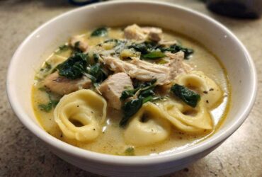 A close-up of a white bowl filled with creamy tortellini soup, featuring chunks of chicken, wilted spinach, and grated Parmesan cheese.
