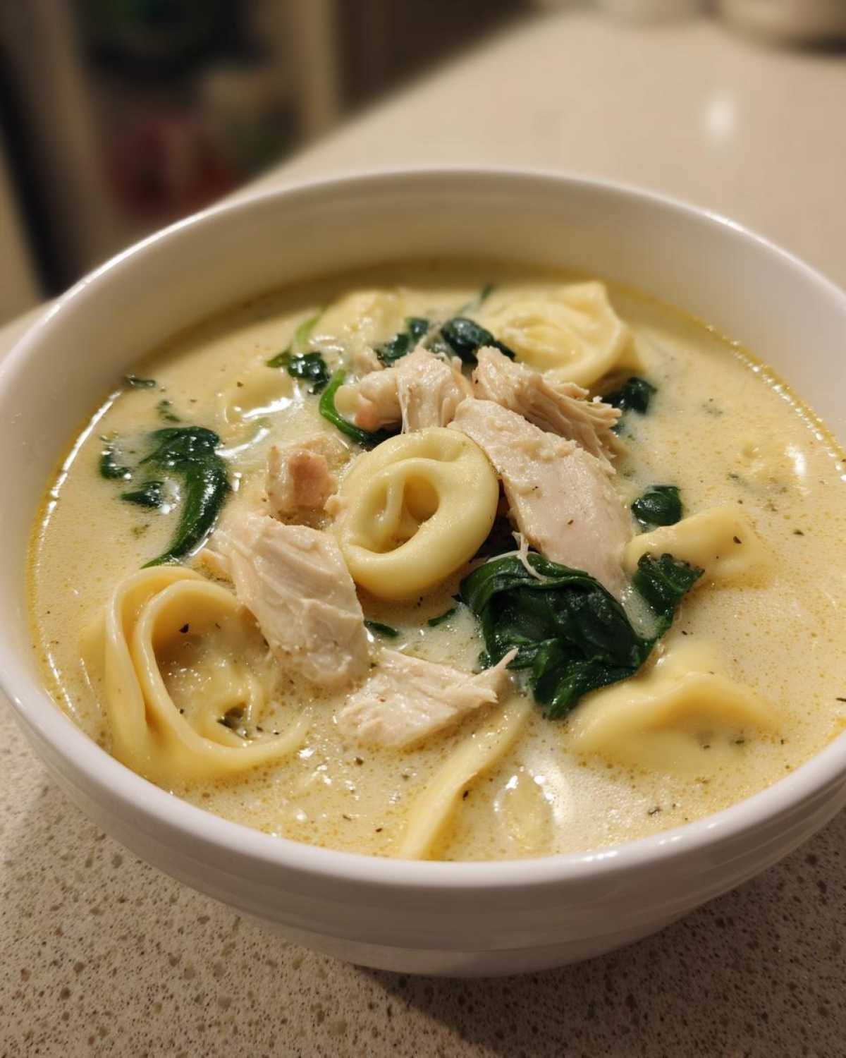 A close-up view of a white bowl filled with creamy tortellini soup, featuring chunks of chicken and wilted spinach.