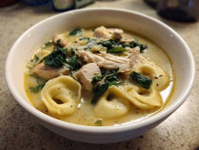 A close-up of a white bowl filled with creamy tortellini soup, featuring chunks of chicken, wilted spinach, and grated Parmesan cheese.