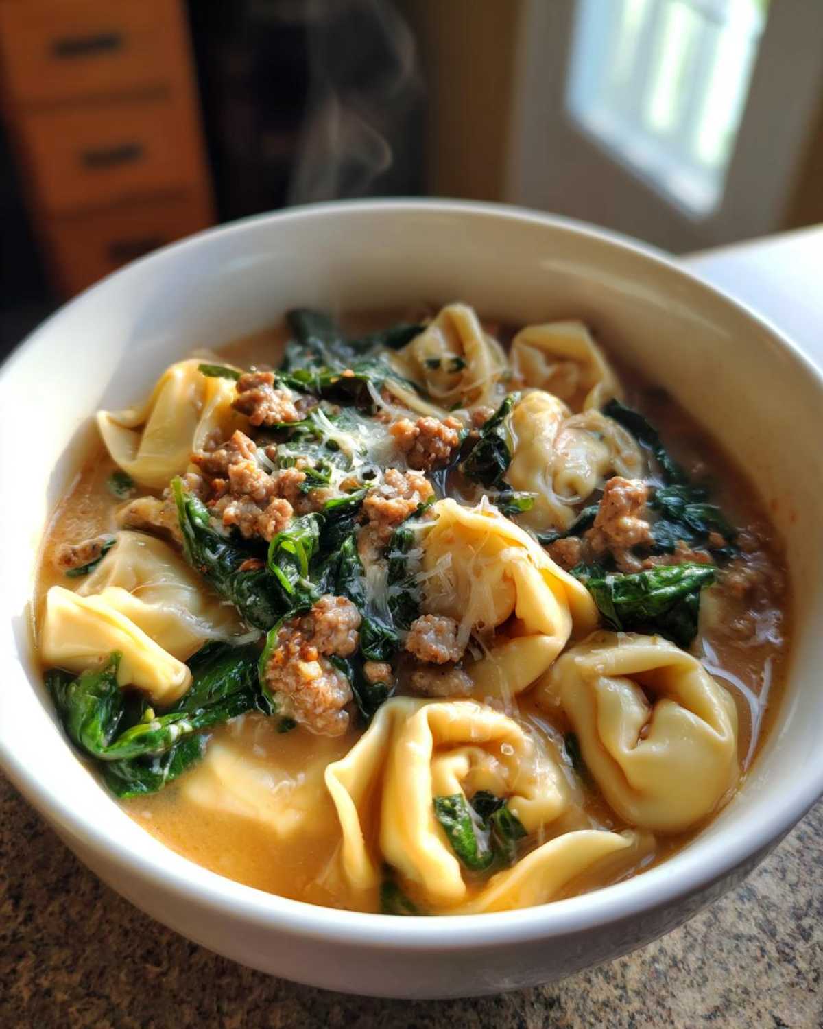 Close-up of a steaming bowl of creamy tortellini soup featuring cheese tortellini, ground sausage, wilted spinach, and grated Parmesan.