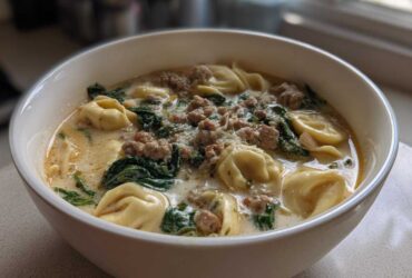 Close-up of a white bowl filled with creamy tortellini soup, featuring sausage crumbles and wilted spinach.