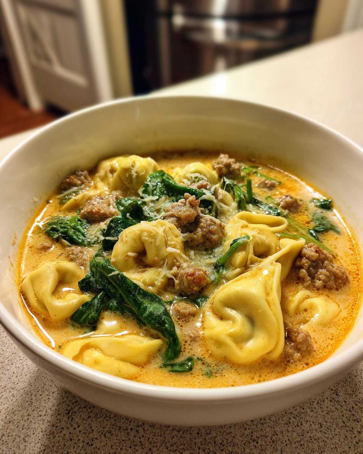 A close-up of a white bowl filled with creamy tortellini soup, featuring cheese tortellini, crumbled sausage, and wilted spinach.