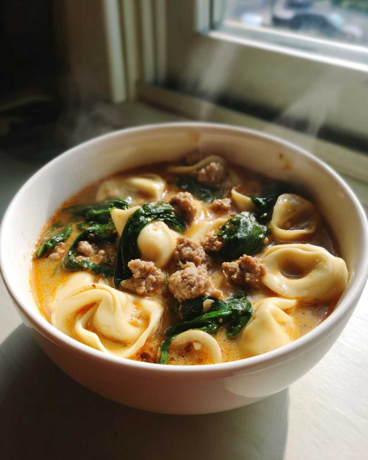 Close-up of a steaming white bowl filled with creamy tortellini soup, featuring cheese tortellini, crumbled sausage, and wilted spinach.