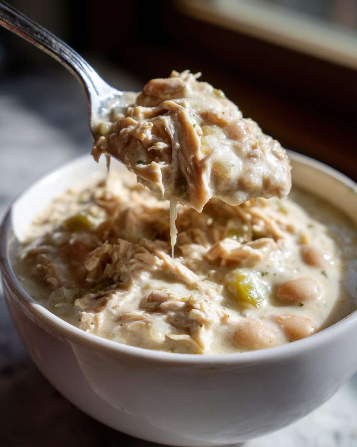 A spoonful of creamy white chicken chili, showing shredded white chicken, beans, and green peppers, lifted from a white bowl.