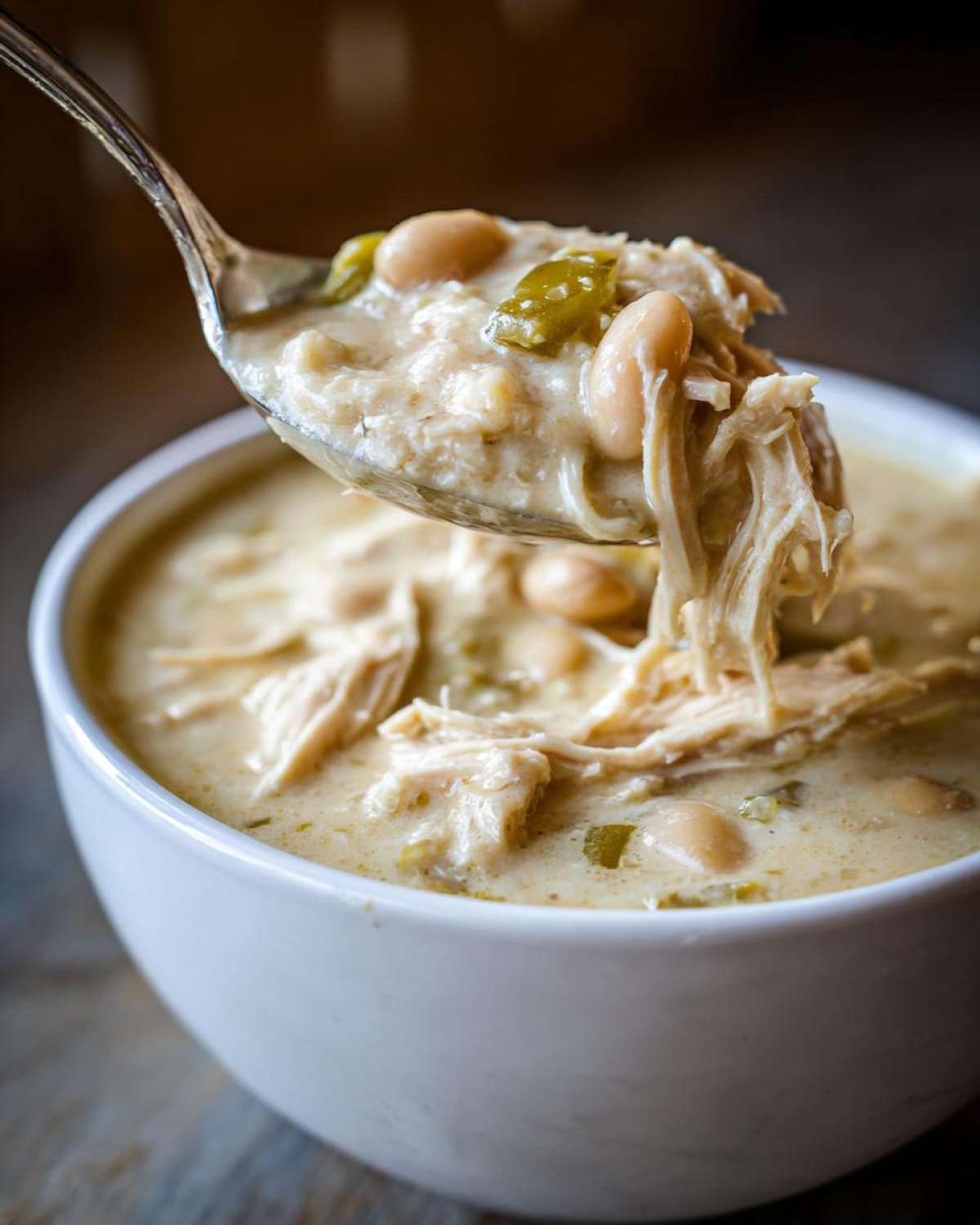 A spoonful of creamy white chicken chili, rich with shredded white chicken, white beans, and green chilies, lifted from a white bowl.