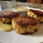 Three golden-brown, pan-fried salmon cakes served with a side of creamy dill sauce.