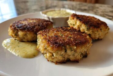 Three golden-brown, pan-fried salmon cakes served with a side of creamy dill sauce.