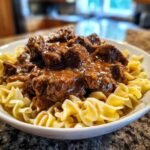Close-up of tender crockpot beef tips smothered in rich brown gravy served over wide egg noodles in a white bowl.