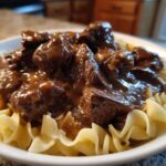 Close-up of tender crockpot beef tips smothered in rich brown gravy served over wide egg noodles in a white bowl.