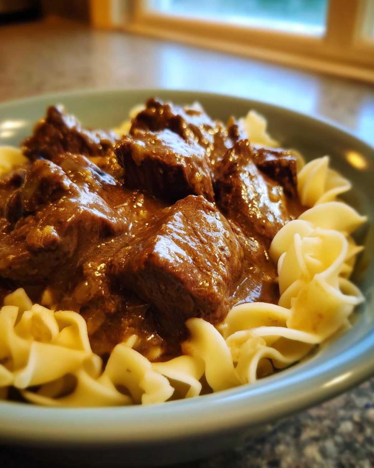 Close-up of tender crockpot beef tips smothered in rich brown gravy served over wide egg noodles in a light blue bowl.