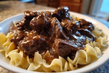Close-up of tender crockpot beef tips smothered in rich brown gravy served over wide egg noodles in a white bowl.