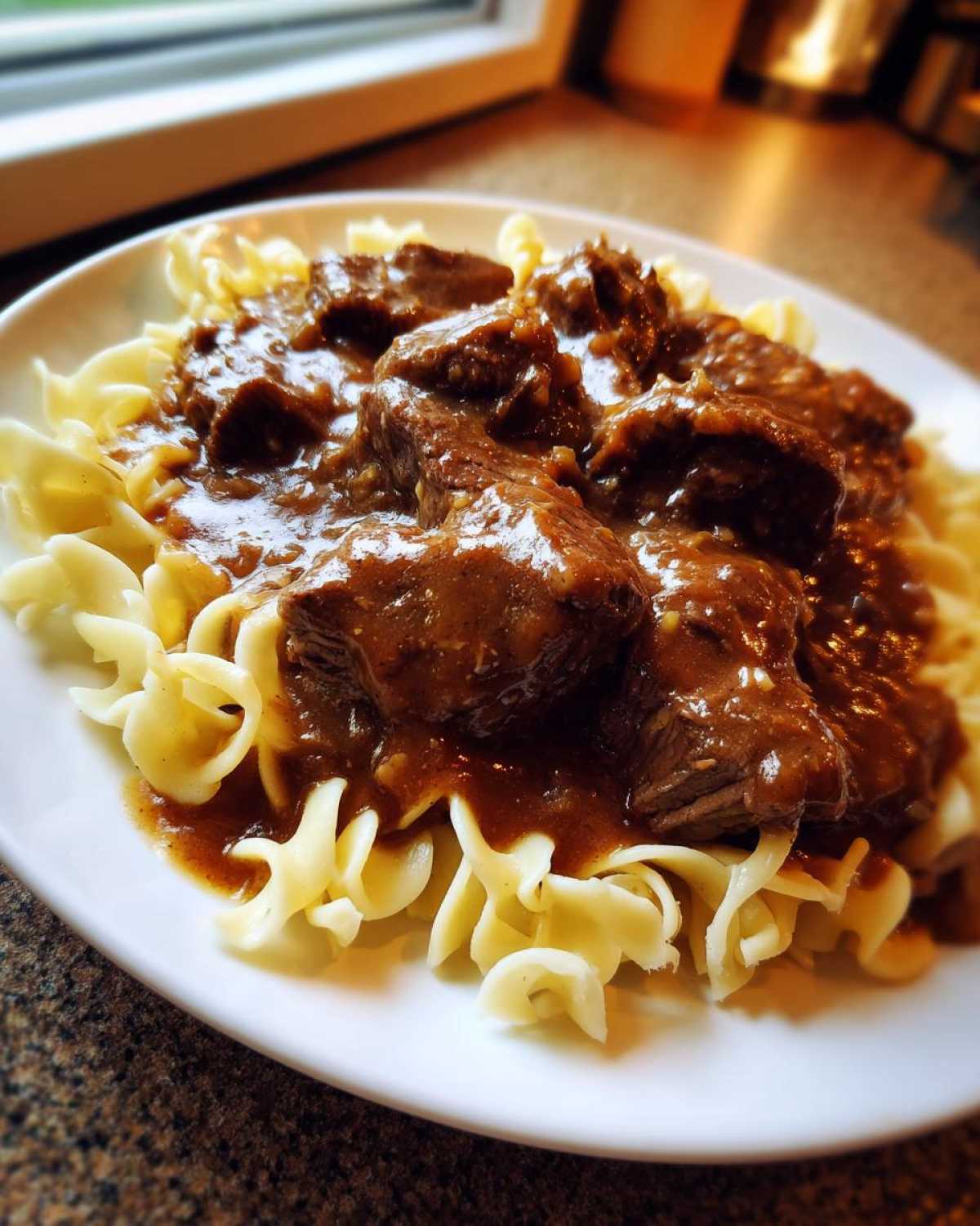 Close-up of tender crockpot beef tips smothered in rich brown gravy served over wide egg noodles on a white plate.